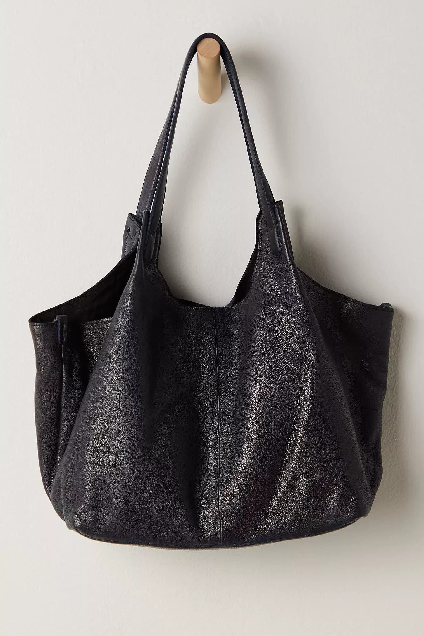 A soft black leather shoulder tote hanging on a wooden peg against a pale wall